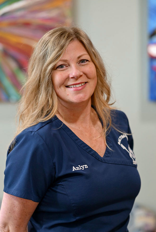The image is a photograph of a woman standing indoors, smiling at the camera. She has long blonde hair and is wearing a dark-colored top with a name tag that includes her first name.