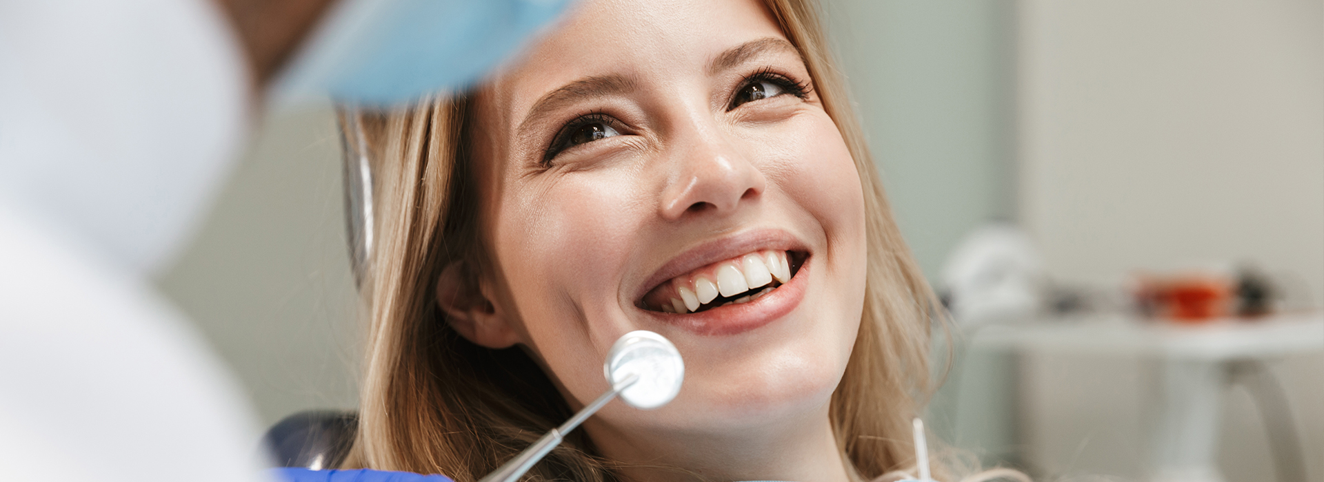 In the image, a woman is smiling at the camera while sitting in a dental chair with a dentist standing nearby. The setting appears to be a dental office, and the woman seems relaxed and comfortable during her visit.