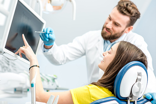 The image shows a dental professional standing next to a patient in a dental office, with the professional pointing at a computer screen.