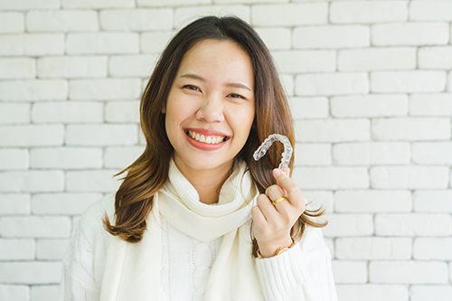 The image shows a smiling woman holding up what appears to be a toothbrush, with her teeth visible and a brick wall in the background.